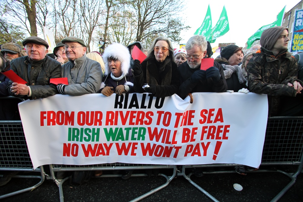 View of protestors, including Activist Maura Harrington (in sunglasses), lined up along the edge of one of the road barricades. There is a sign draped over the fencing which reads "Rialto / From our rivers to the sea / Irish Water will be free / No Way We Won't Pay!".