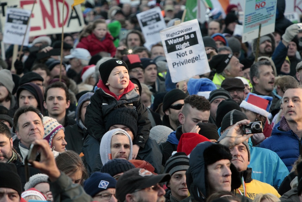 Close-up of the crowd showing a boy seated on top of an adult's shoulders. A printed sign in centre reads "Right2Water / Water is a Right / Not a Privilege!"