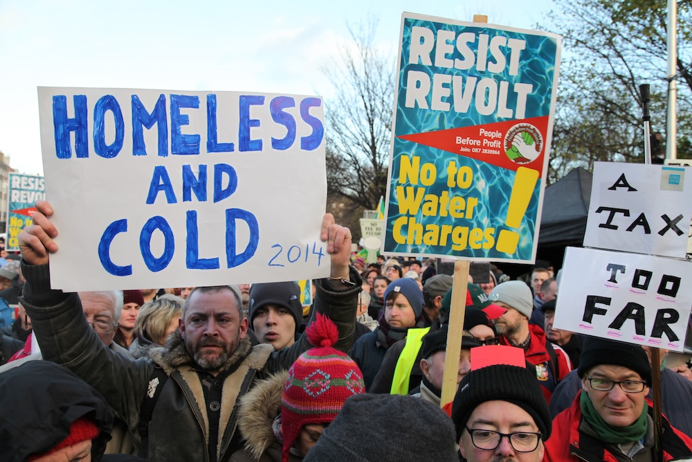 View of a crowd with placards reading "Homeless and Cold", " RESIST REVOLT / People Before Profit / No to Water Charges!" and "A Tax Too Far".