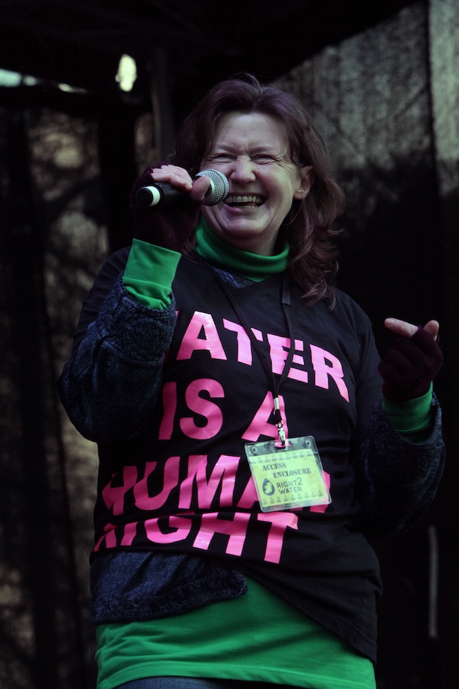 Noreen Murphy, Cork Right2Water, wears a dark blue shirt with pink lettering reading "Water is a Human Right" holds a microphone on stage.