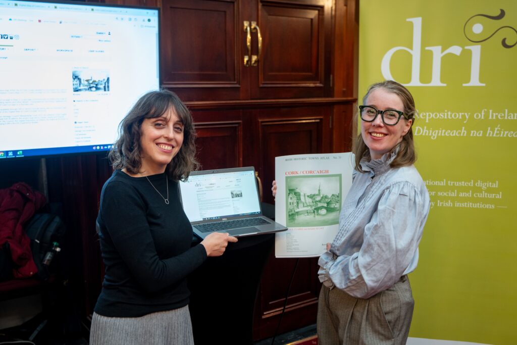 Two people stand indoors; one holds a laptop displaying a webpage, and the other holds a poster. The Digital Repository of Ireland banner is visible in the background.