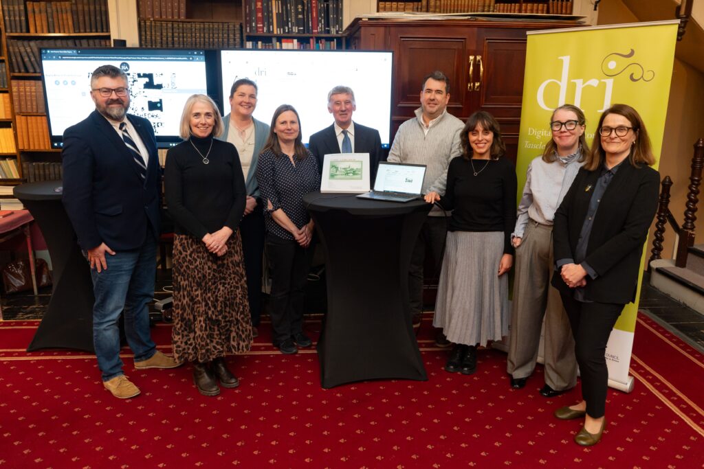 A group of nine people stands together in a room with bookshelves. They are posing in front of two monitors and a banner that reads "DRI."